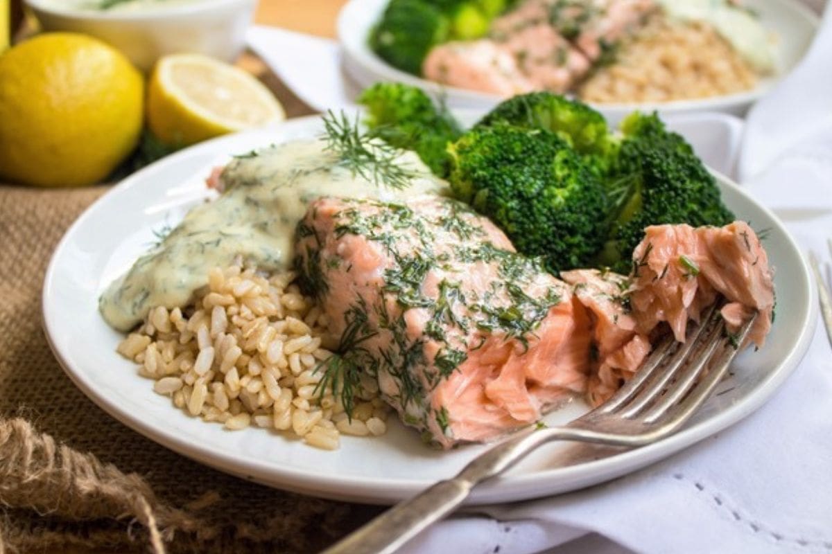 salmon with creamy dill sauce on top next to brown rice and broccoli on a plate