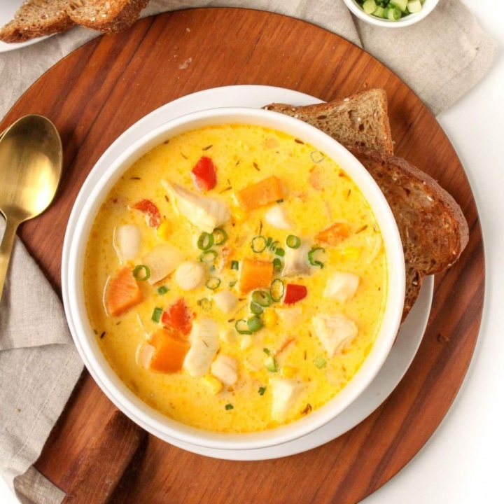 a bowl of seafood chowder with sweet potato on top of a wooden platter with crusty bread beside it