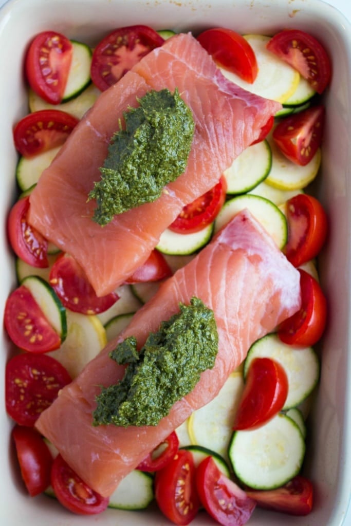raw salmon filets with pesto sauce on top of sliced zucchini and tomatoes in a baking dish