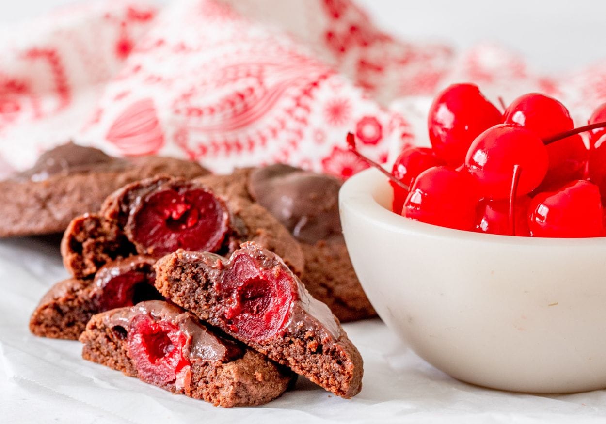 A pile of chocolate covered cherry cookies cut in half next to a marble bowl with maraschino cherries.