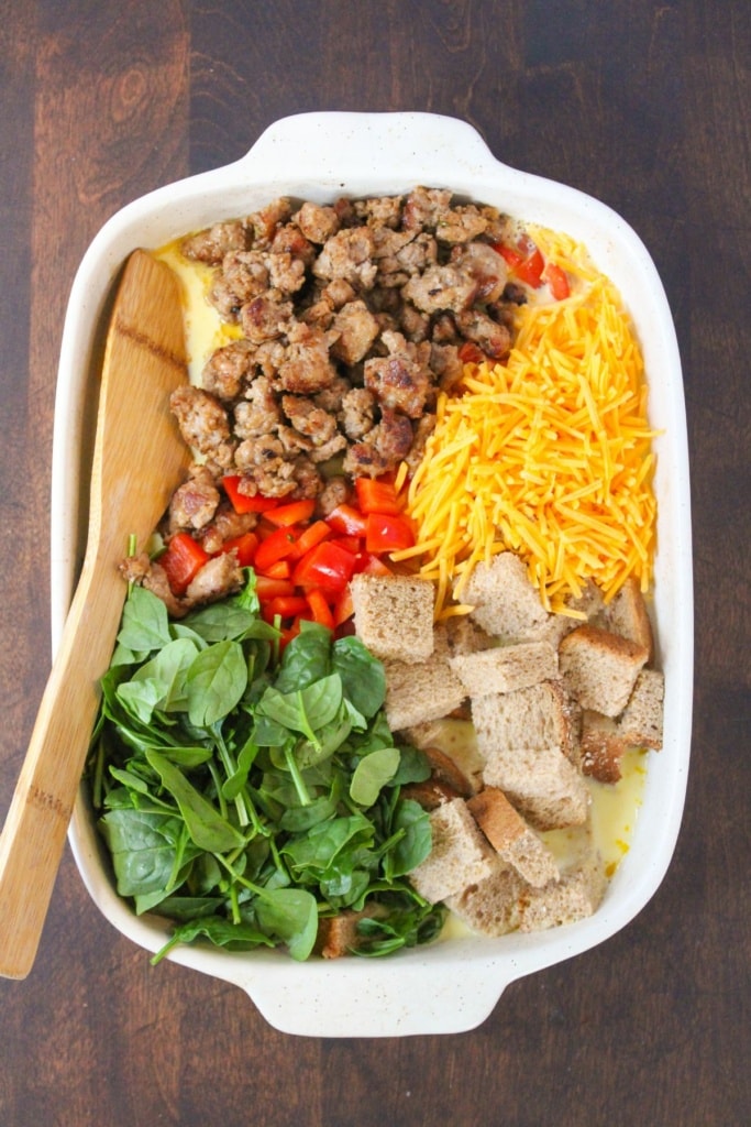 a white baking dish with cooked sausage, spinach, shredded cheddar cheese, bread cubes, and diced bell peppers with a wooden spoon on top