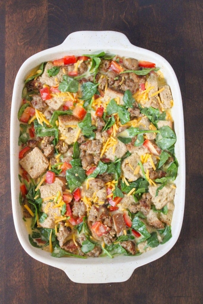 a white baking dish with sausage, diced vegetables, bread cubes, and shredded cheese stirred together