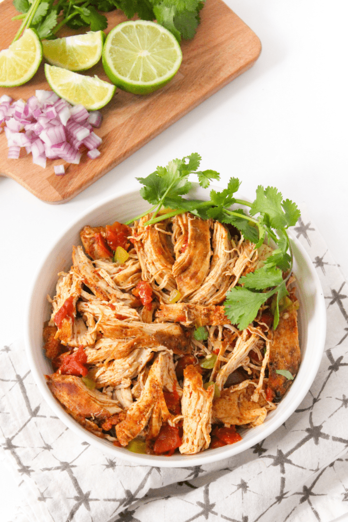 crockpot shredded chicken in a bowl with sliced limes on a wooden platter behind it