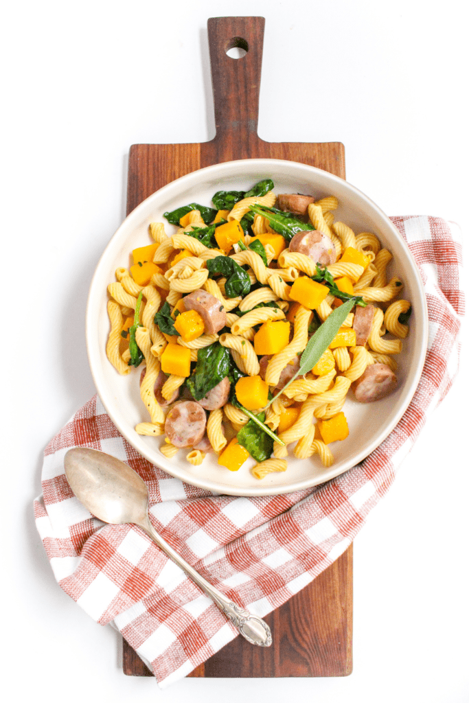 chicken sausage and chickpea spirals with veggies in a white bowl with a checkered towel and wooden platter underneath