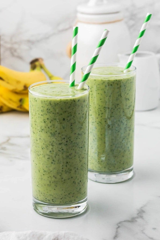 two green smoothies in tall glasses with striped straws sitting on a marble countertop