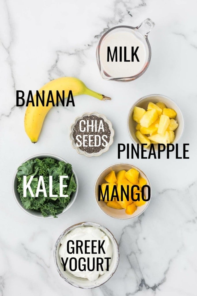 pineapple, mango, banana, kale, Greek yogurt, milk, and chia seeds in small bowls on a marble countertop