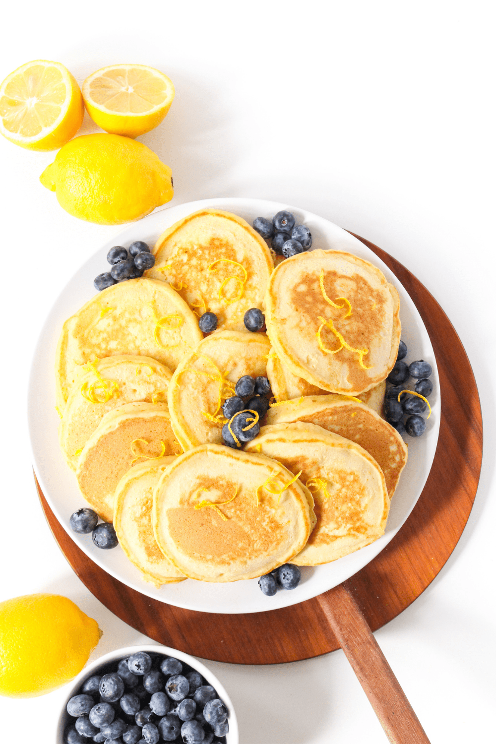 lemon pancakes with ricotta spread out on a plate, with lemon slices and a bowl of blueberries beside it
