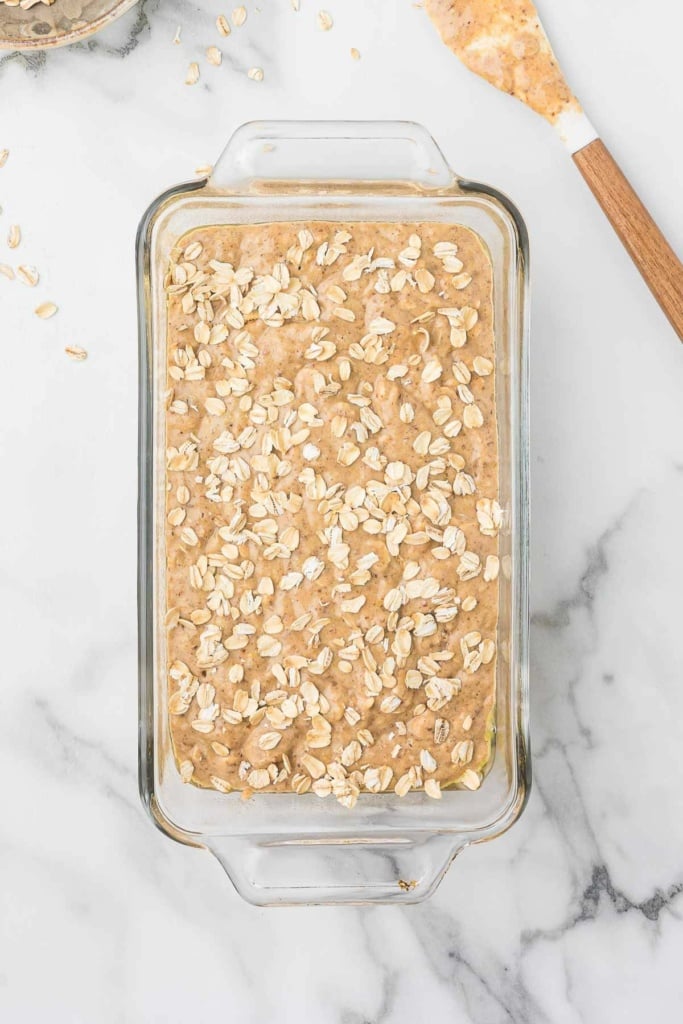 whole wheat banana bread batter in a glass loaf pan sprinkled with oats on top