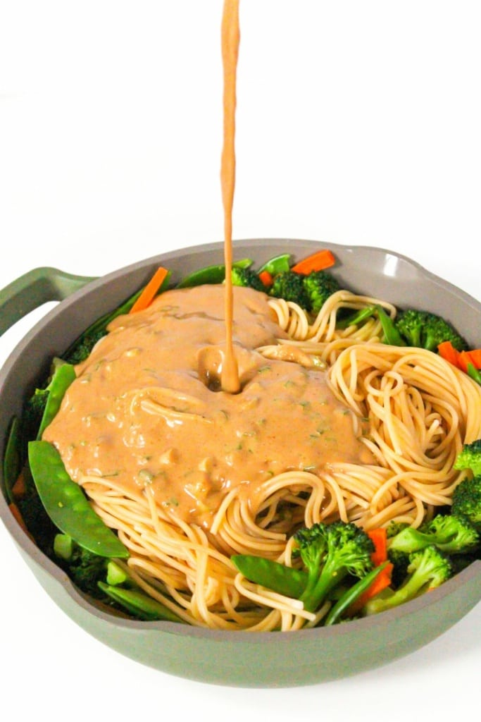 peanut sauce being poured over a large skillet with noodles and vegetables