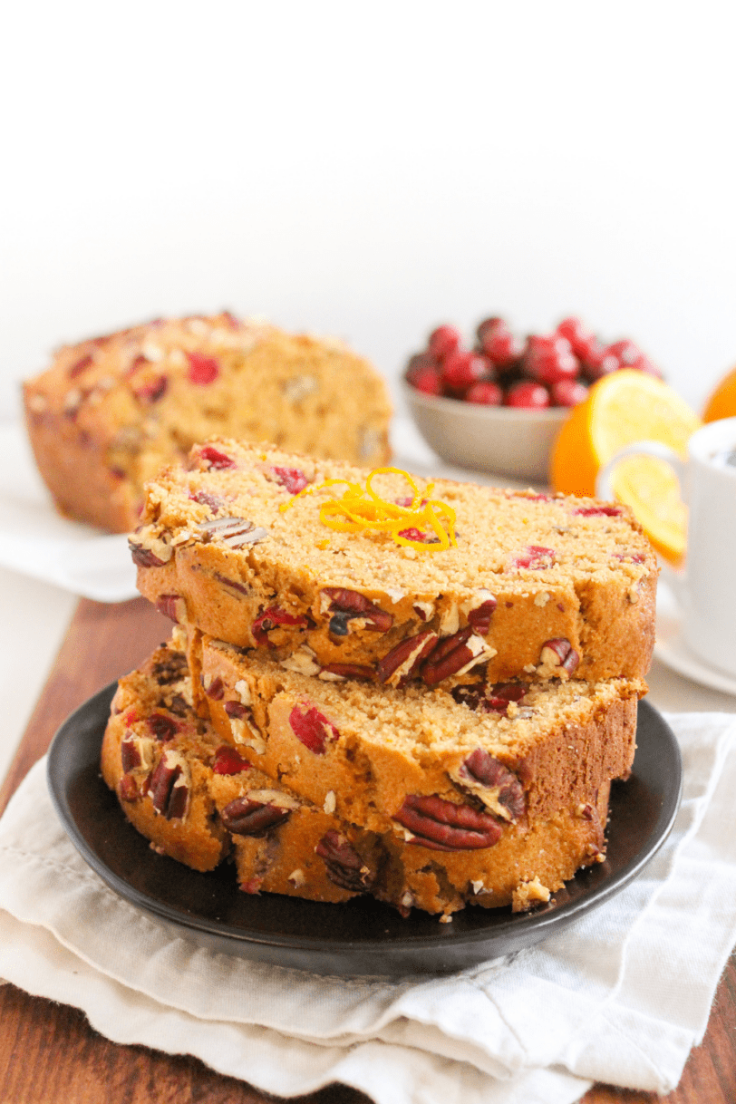slices of cranberry orange bread stacked on a black plate with a bowl of cranberries in the background