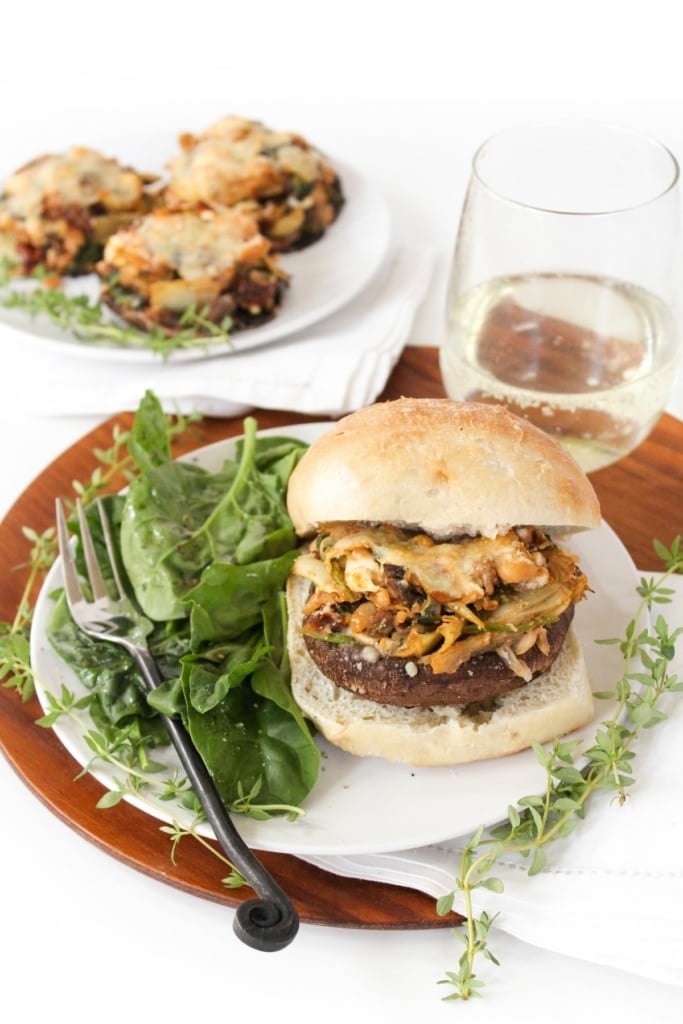 A stuffed portobello mushroom inside a bun with a green salad on the side, with a stemless glass of white wine behind it.