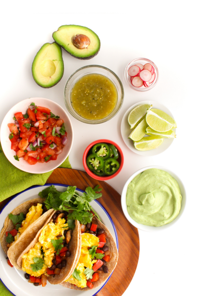 breakfast tacos with small bowls of toppings: avocado crema, green salsa, pico de gallo, limes, radishes, and jalapeno slices