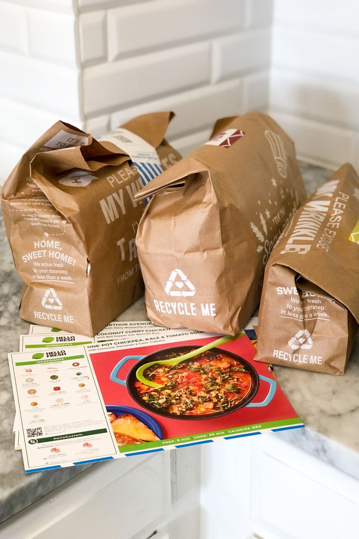 HelloFresh recipe cards on a marble counter next to HelloFresh paper bags with ingredients inside