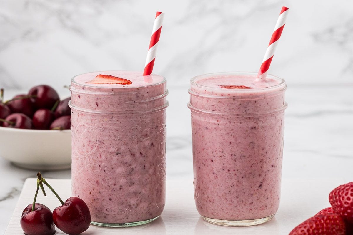 two strawberry cherry smoothies in mason jars with striped red and white straws on a marble countertop