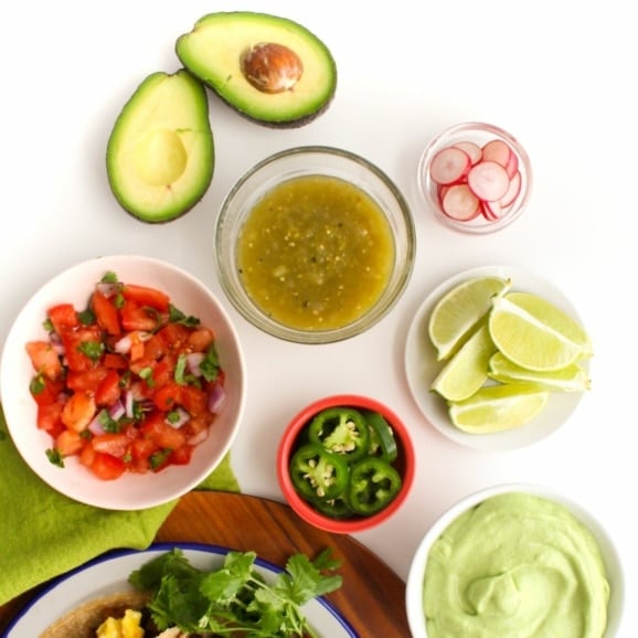 taco toppings in small bowls, including salsa verde, pico de gallo, avocado crema, sliced jalapenos, and more