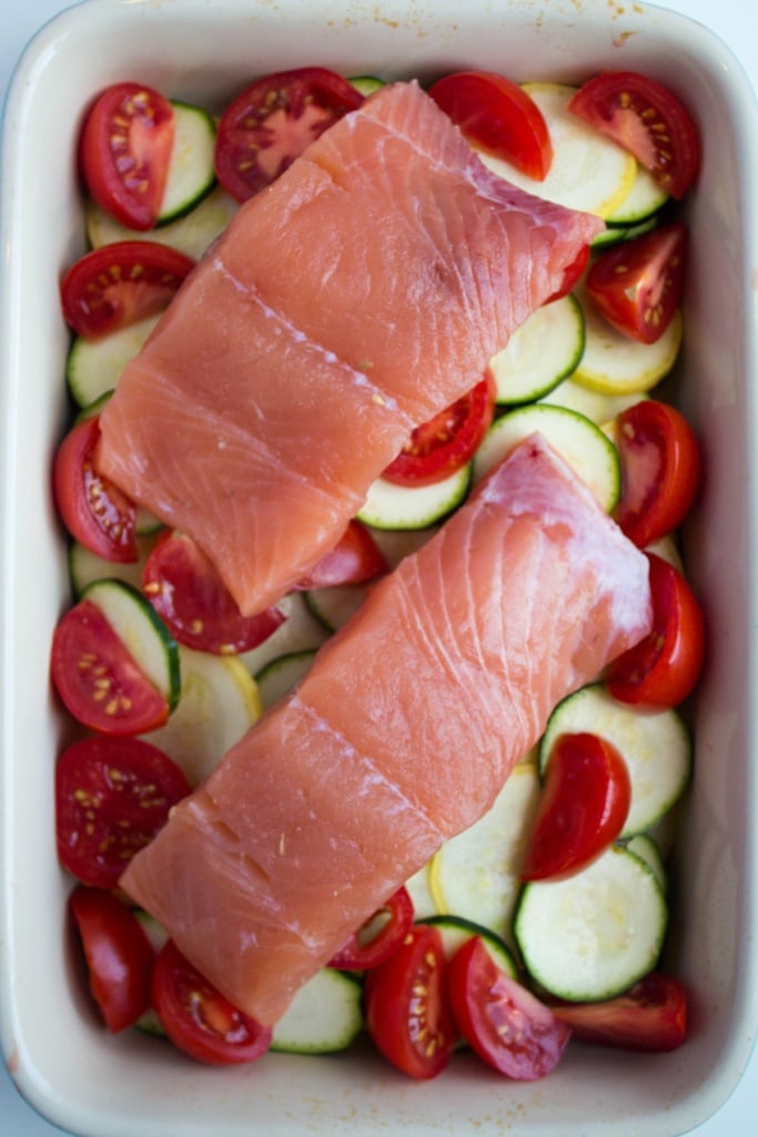 raw salmon filets on top of sliced zucchini and tomatoes in a baking dish