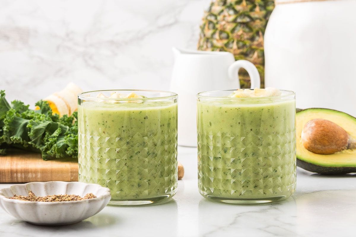 two green avocado smoothies in short glasses with kale and pineapple in the background