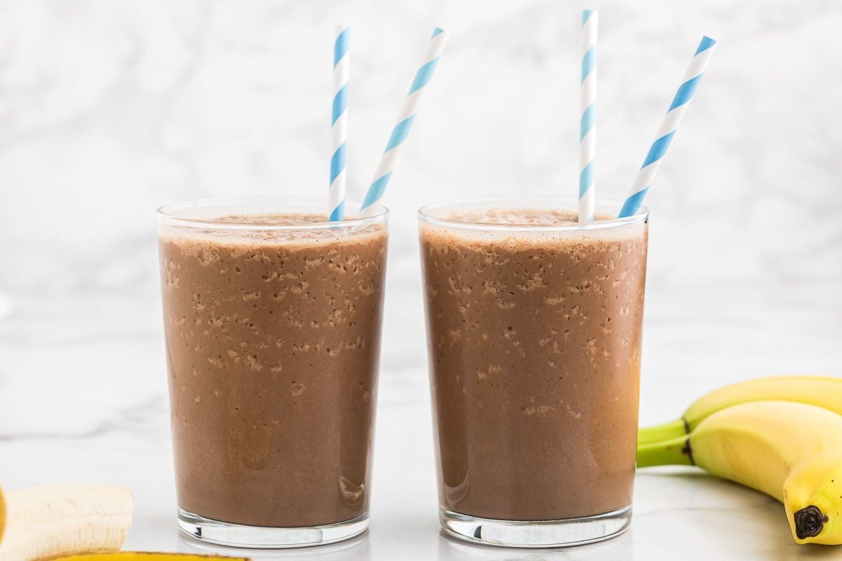 two chocolate smoothies in short glasses with bananas next to them