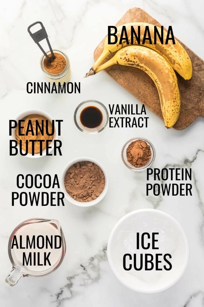 bananas, peanut butter, almond milk, protein powder, cocoa powder, cinnamon, vanilla extract, and ice cubes in small bowls on a white marble surface