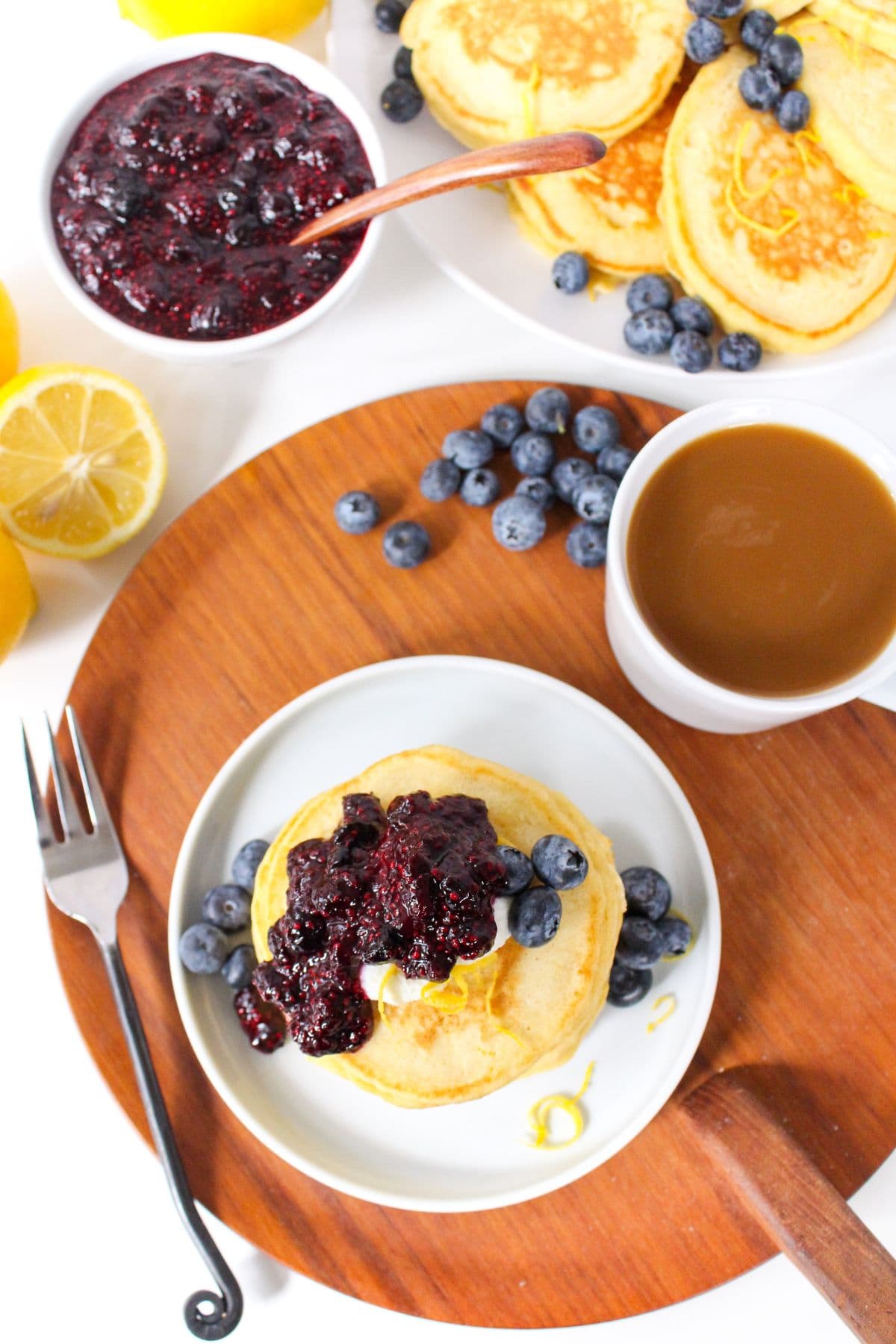 a bowl of blueberry chia jam next to a stack of lemon pancakes on a white plate and a mug of coffee on a wooden platter