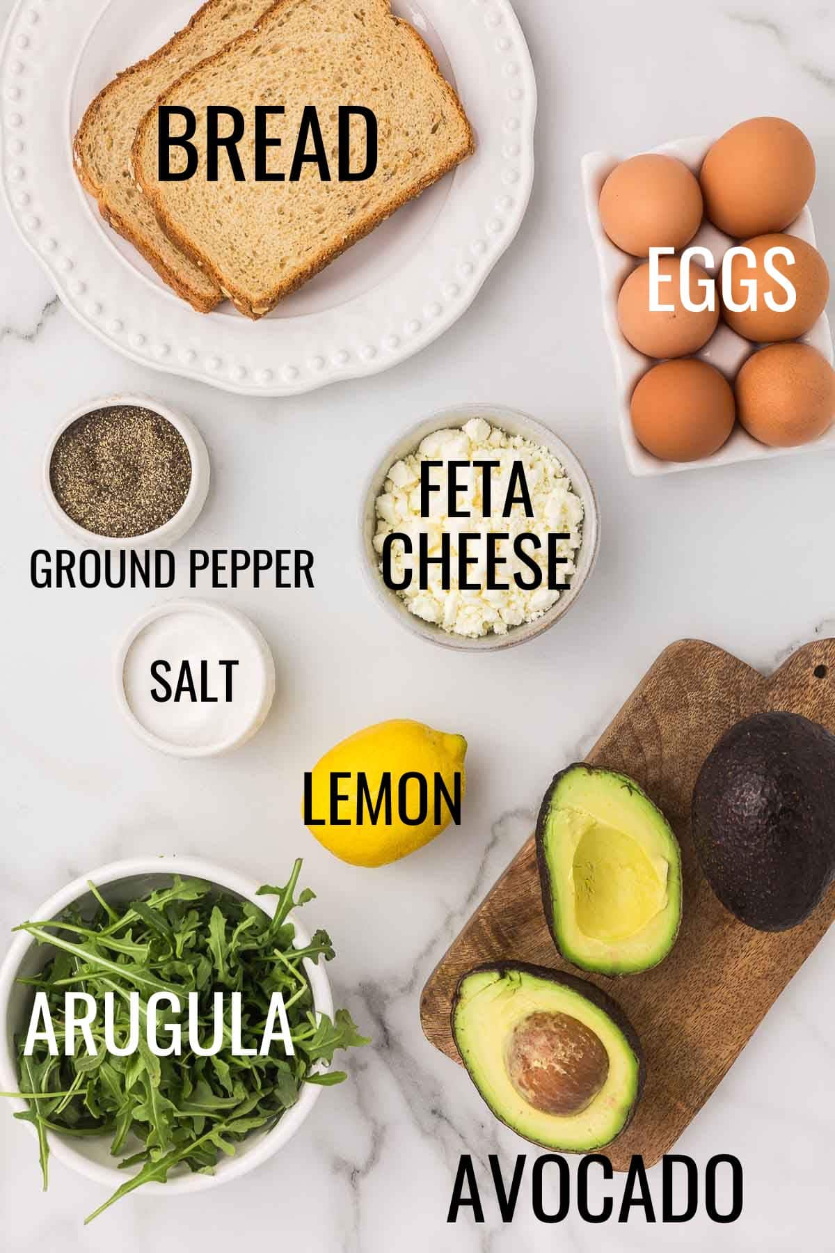 feta cheese, eggs, bread, arugula, avocado, lemon, salt, and pepper in small bowls on a marble countertop