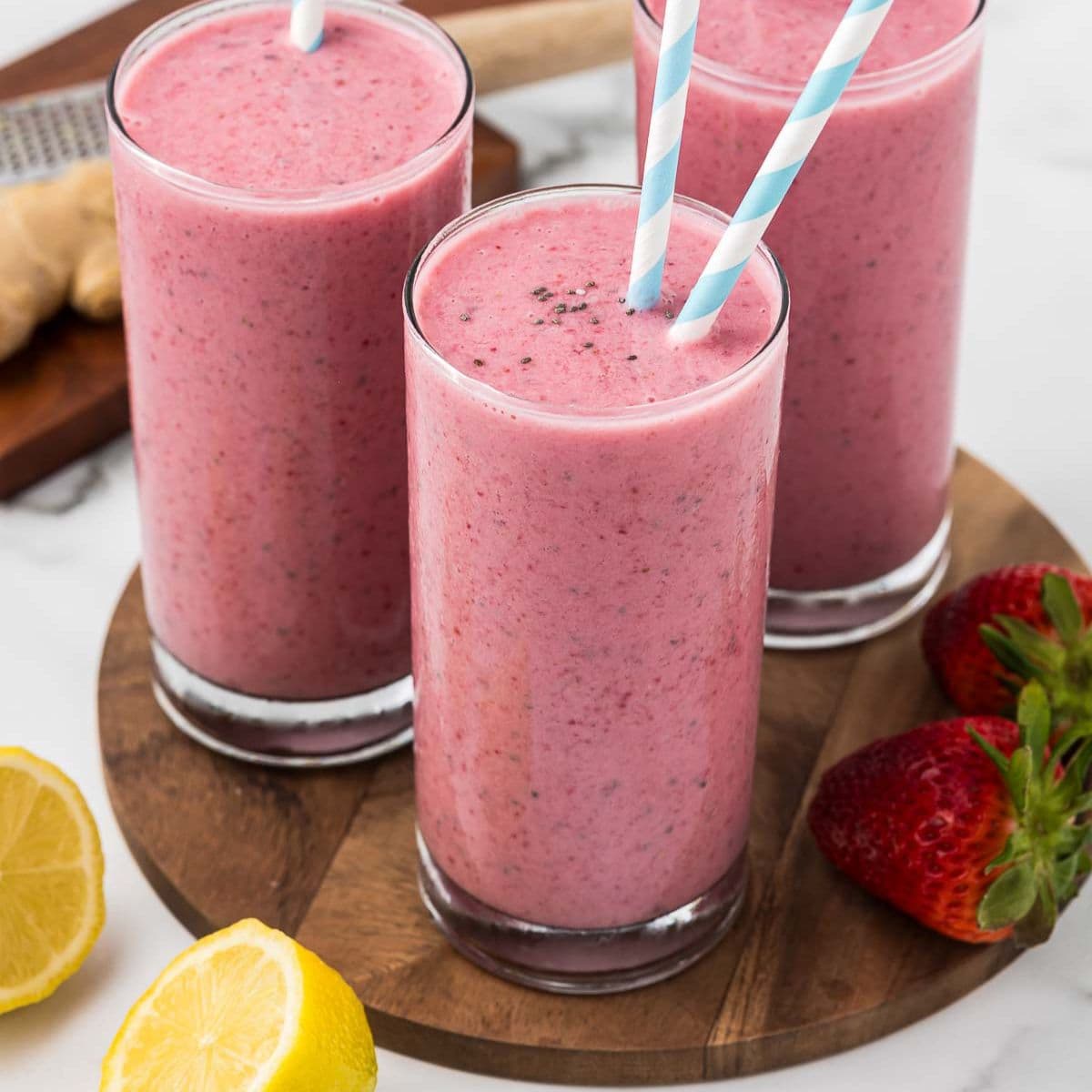 three pink beet fruit smoothies in tall glasses with striped straws on a wooden platter