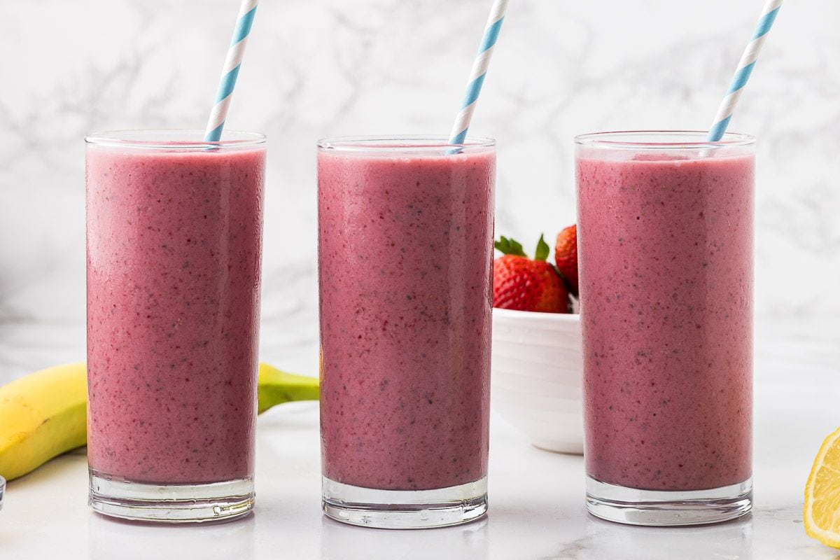 three pink beet fruit smoothies with blue and white striped straws next to a banana on a white marble countertop